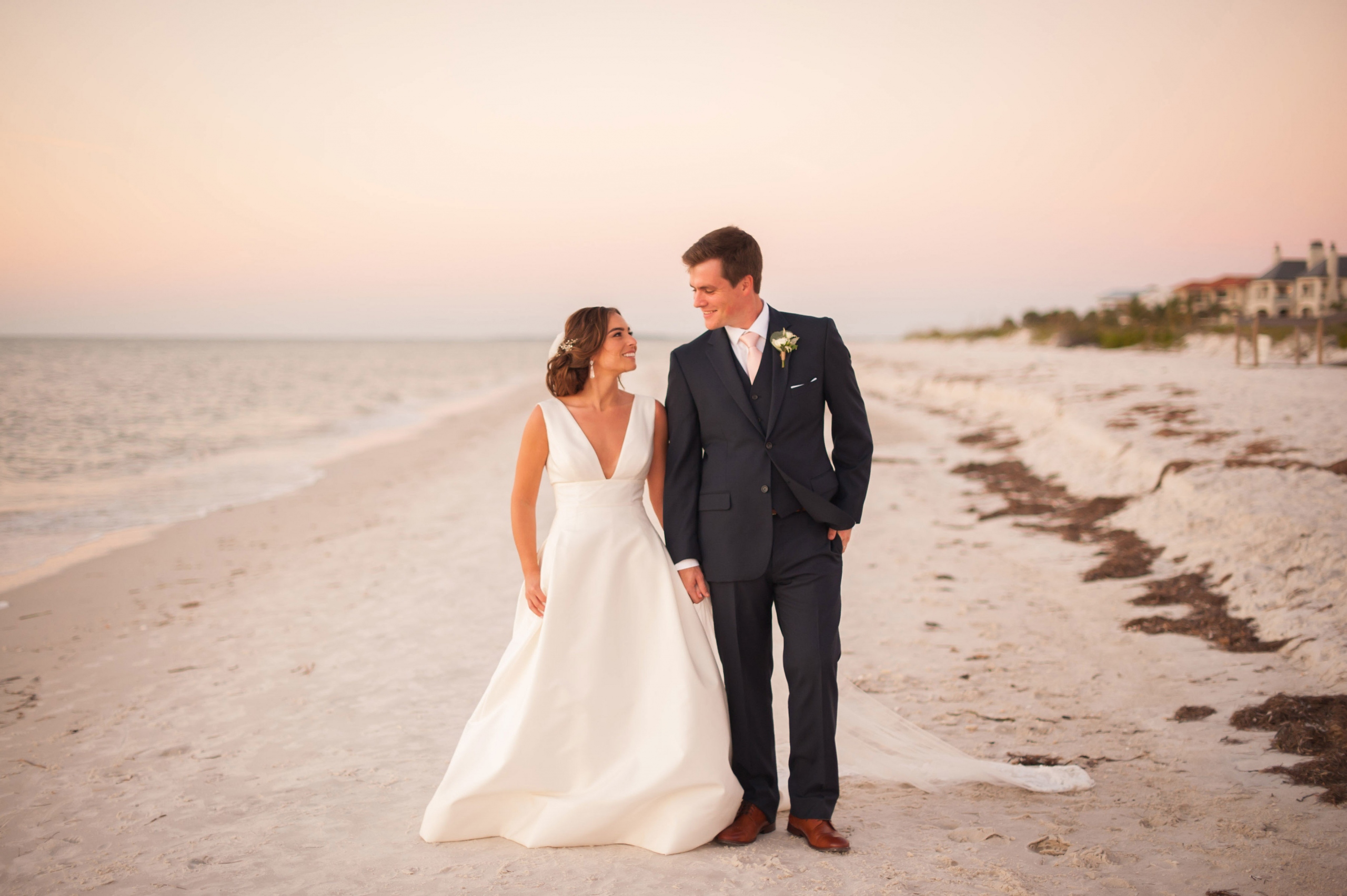 a bride and groom standing on the beach at sunset